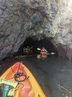 View of the tip of an orange kayak in the water in the foreground with the opening of a cave in the background.